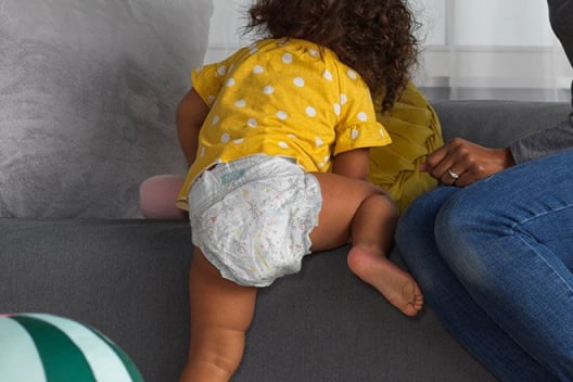 A baby wearing a Huggies Little Movers Plus Diaper climbs onto a couch next to their mother