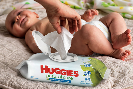 A smiling baby lays on a blanket while a hand pulls a wipe from a package of Huggies Natural Care Refreshing Wipes with EZ Pull lid