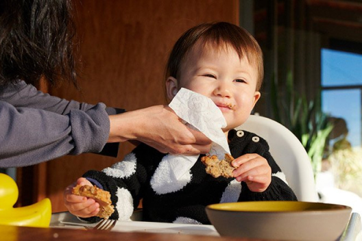 A mother leans down to wipe her baby's messy face while they eat in a high chair